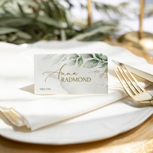May include: A white place card with a watercolor design of green leaves and gold accents. The text "Anna Radmond" is written in gold script font. The text "Table One" is printed in gold at the bottom of the card.
