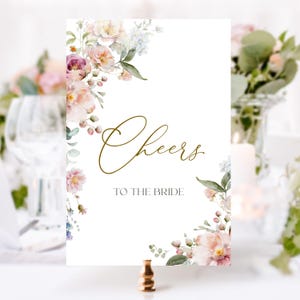 May include: A white card with a floral design in shades of pink, blue, and green. The card says "Cheers to the Bride" in gold lettering.