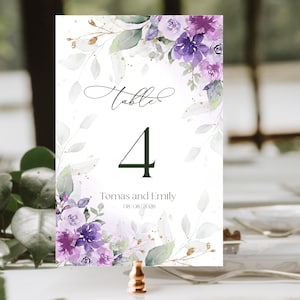 May include: A white table number card with floral watercolor design. The card features the word "Table" in script, the number "4", and the names "Tomas and Emily" with a date. Purple and green flowers and leaves decorate the edges.