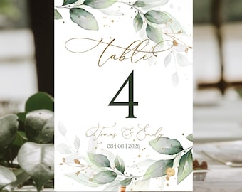 Op de afbeelding: Een witte tafelnummerkaart met groene en gouden aquarelbladeren en gouden glitter. De kaart heeft de tekst "Tafel 4" en "Tomas & Emily 08 01 08 | 2026".
