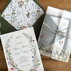 May include: Wedding invitation suite featuring a floral wreath design with the names Stephen Hudson and Olivia Martin. The invitation includes the wedding date, time, and location. The suite includes an envelope with a floral liner and vellum wrap tied with twine.