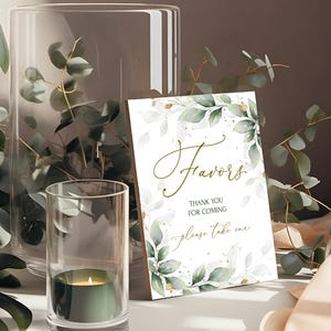 Puede incluir: Un cartel blanco con letras doradas que dice "Favors" y "Gracias por venir - por favor, toma uno". El cartel está decorado con un diseño de acuarela de hojas verdes y purpurina dorada.