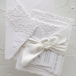 May include: A white stationery set featuring deckled edges and embossed floral patterns on the envelope flap. The set includes a response card with text reading ACCEPTS WITH PLEASURE and DECLINES WITH REGRET, tied together with a white ribbon and a wax seal.