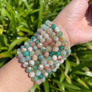 Puede incluir: Una pila de pulseras de cuentas de ágata verde, blanca y rosa. Las pulseras están hechas de cuentas redondas y están apiladas en la muñeca de una persona.