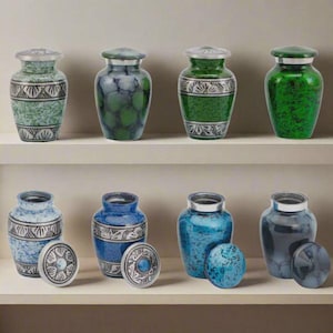 May include: A selection of decorative urns in shades of green, blue, and grey. Each urn has a silver-coloured rim and lid. Some urns feature patterned designs, while others have a speckled or textured appearance.