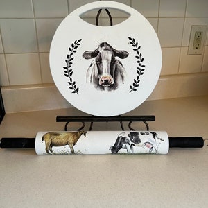 May include: A white round cutting board with a black and white cow design and a black wreath. A rolling pin with a white background and black and white cow designs.