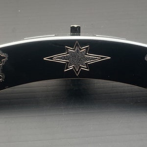 May include: A curved, black decorative object with a silver starburst design in the center. The ends feature detailed silver oak leaf and acorn patterns. The object has a small, raised silver piece on top.