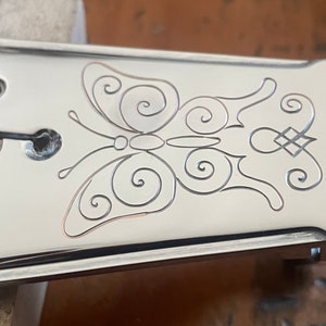 Pode incluir: Uma placa de ponte de guitarra de metal prateado com um design decorativo de borboleta. A placa tem quatro furos para parafusos e um pequeno parafuso de ajuste no lado direito.