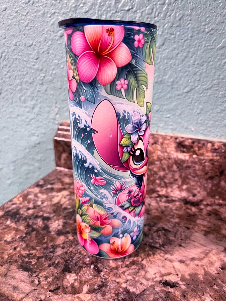 May include: A pink and blue stainless steel tumbler with a floral design and a cartoon character of Stitch from Lilo & Stitch riding a wave. The tumbler has a blue lid.