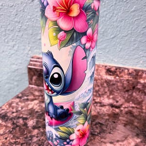 May include: A blue and pink stainless steel tumbler with a cartoon image of Stitch from Lilo & Stitch surfing on a pink wave. The tumbler is surrounded by tropical flowers and leaves.