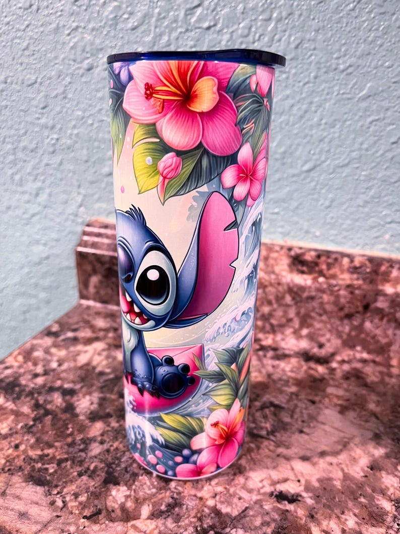 May include: A blue and pink stainless steel tumbler with a cartoon image of Stitch from Lilo & Stitch surfing on a pink wave. The tumbler is surrounded by tropical flowers and leaves.