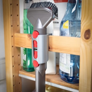 May include: A gray vacuum cleaner attachment with a red button and a brush on the end. The attachment is mounted on a wooden shelf.