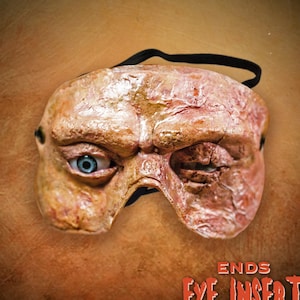 以下が含まれることがあります： 縫い付けられた目と、詳細な質感のある肌色のデザインが施された、リアルな目のマスク。マスクには黒いゴムバンドが付いています。テキスト「ENDS EYE INSERT」は、白地に赤で印刷されています。