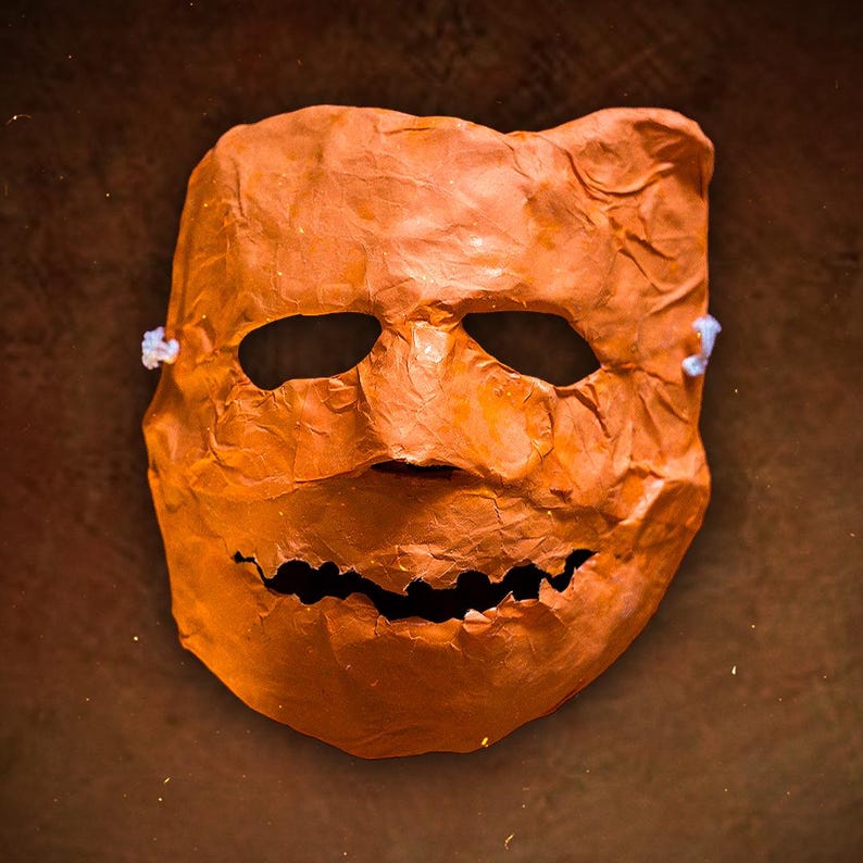 May include: A bright orange paper mache mask with a wide, toothy grin. The mask has a wrinkled texture and is designed to look like a human face.
