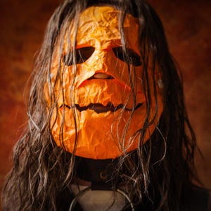 May include: A person wearing a bright orange pumpkin mask with a carved smile. The mask is made of paper and has a wrinkled texture. The person's long brown hair is covering most of the mask.