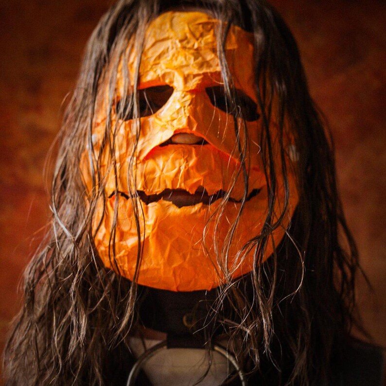 May include: A person wearing a bright orange pumpkin mask with a carved smile. The mask is made of paper and has a wrinkled texture. The person's long brown hair is covering most of the mask.