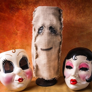 Puede incluir: Tres máscaras de Halloween sobre una superficie marrón. Las máscaras incluyen una máscara de saco de arpillera con ojos cosidos y una sonrisa, y dos máscaras de porcelana con rasgos pintados, incluyendo labios rojos y mejillas rosadas. El fondo es un degradado de naranja y marrón.