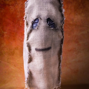 Puede incluir: Una máscara de saco de yute con agujeros para los ojos y una sonrisa pintada. La máscara está montada sobre un soporte negro, con una apariencia áspera y texturizada. El fondo es un degradado de naranja y marrón.