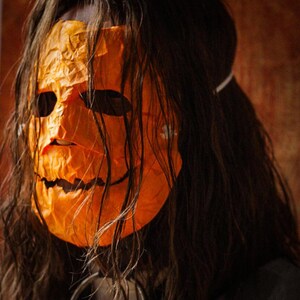 May include: A person wearing a long, dark brown wig and an orange pumpkin mask with a wide, toothy grin.