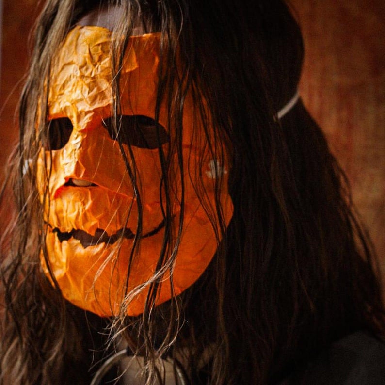 May include: A person wearing a long, dark brown wig and an orange pumpkin mask with a wide, toothy grin.