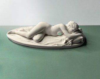 May include: A white porcelain sculpture of a reclining nude figure, lying on a pillow with a horn of plenty at their feet.