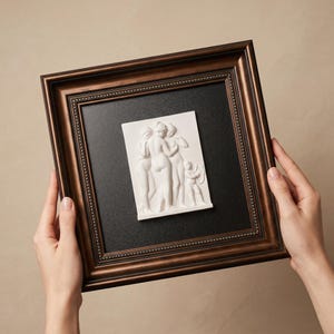 May include: A framed white relief sculpture of three figures and a child. The artwork is set against a black background within a brown, ornate frame. The frame is held by two hands against a neutral background.