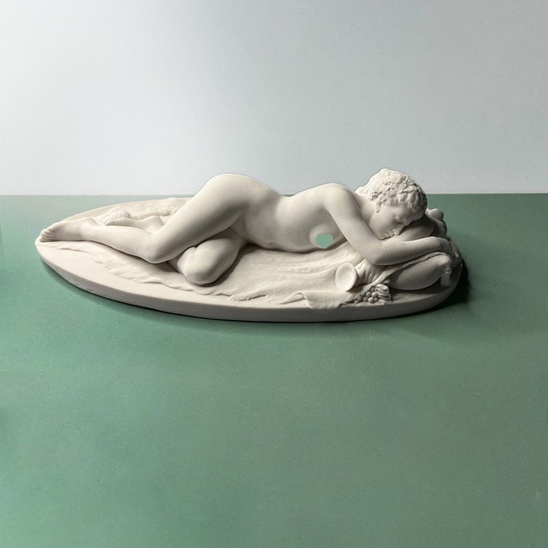May include: A white porcelain sculpture of a reclining nude figure, lying on a pillow with a horn of plenty at their feet.