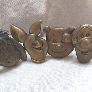 May include: A set of four brass-toned metal cabinet knobs featuring the sculpted faces of Eeyore, Piglet, Winnie the Pooh, and Tigger. Each knob has a detailed relief design with a metallic finish and a central mounting post on the back.