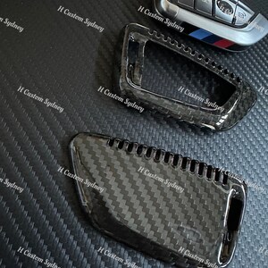 May include: Two black carbon fiber key covers for a car key. The covers are textured and have a glossy finish. The key covers are designed to fit over the existing key fob.