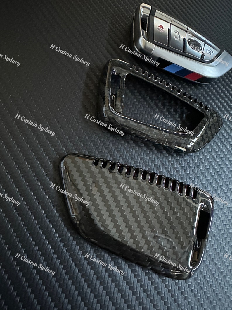 May include: Two black carbon fiber key covers for a car key. The covers are textured and have a glossy finish. The key covers are designed to fit over the existing key fob.