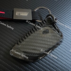 May include: A black carbon fiber key fob cover with a silver key ring. The cover has a textured surface and the text "EWIII" is printed on the cover.
