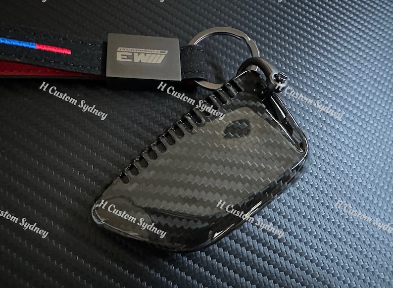 May include: A black carbon fiber key fob cover with a silver key ring. The cover has a textured surface and the text "EWIII" is printed on the cover.