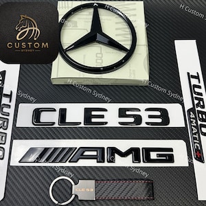 May include: A collection of black automotive emblems including a Mercedes-Benz star logo, CLE 53 and AMG lettering, and two Turbo 4MATIC+ badges. Also included is a black carbon fiber style keychain with the text CLE 53 and a logo square.