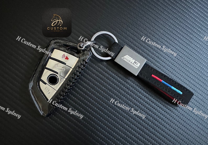 May include: A black carbon fiber key fob with a silver key and a black leather keychain with a silver M3 logo and red, blue, and white stitching.
