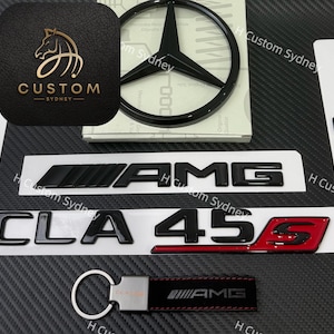 May include: A collection of black car emblems and accessories, including AMG and CLA 45 S badges, a Mercedes-Benz emblem, and a keychain. The items are displayed on a dark, textured surface.