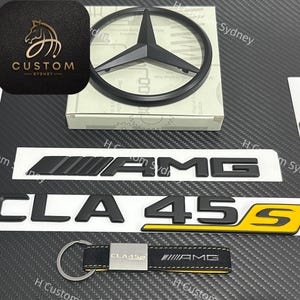 May include: A collection of black car emblems and accessories, including a Mercedes-Benz emblem, AMG badges, and a CLA 45 S badge with yellow accents. Also includes a keychain with AMG branding. The items are displayed on a carbon fiber textured surface.