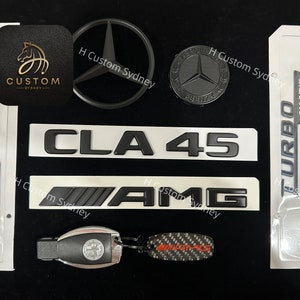 May include: A set of black and silver car emblems with the words "TURBO 4MATIC", "CLA 45", and "AMG". There is also a black Mercedes-Benz star emblem and a black key fob with a silver Mercedes-Benz star emblem.