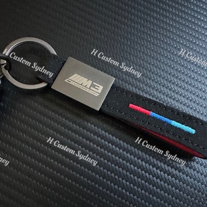 May include: A black leather keychain with a silver metal tag that says "M3 COMPETITION". The keychain has a red, blue, and white stripe and the text "H Custom Sydney" is printed on the background.