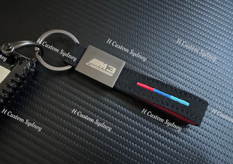 May include: A black leather keychain with a silver metal tag that says "M3 COMPETITION". The keychain has a red, blue, and white stripe and the text "H Custom Sydney" is printed on the background.