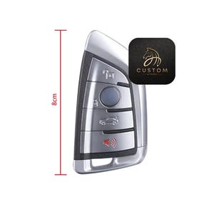 May include: A silver car key fob with five buttons, including a lock, unlock, trunk, and panic button. The key fob is 8 cm long.