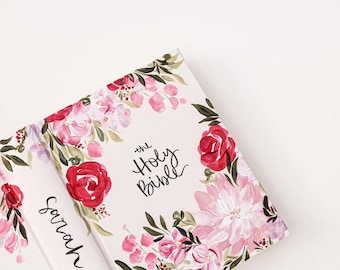 May include: A white floral design covers the front and back of a hardcover book. The front cover features the text "the Holy Bible" in black cursive lettering. The back cover has the name "Sarah" written in black cursive lettering.