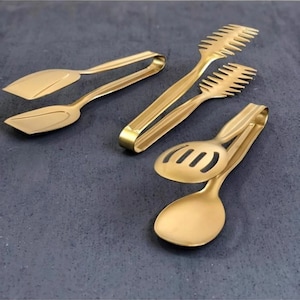 Puede incluir: Conjunto de cuatro tenazas de cocina doradas. Las tenazas incluyen una cuchara ranurada, una espátula y dos pares de tenazas con diseños diferentes.