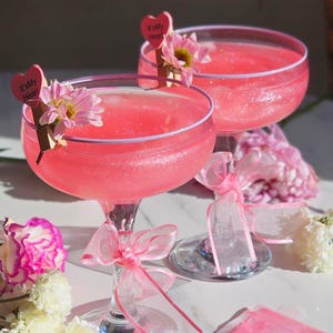 Pode incluir: Dois coquetéis rosa em taças coupe, decorados com flores e etiquetas em forma de coração com a frase "It's My Heart". Cada taça tem uma fita rosa amarrada ao redor da haste. Um pacote de glitter comestível é visível.