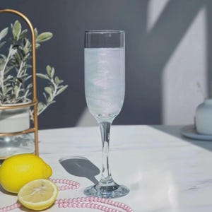 May include: A tall, clear champagne flute filled with a sparkling, pale liquid. A whole yellow lemon and a lemon half sit beside the glass, with a strand of pink beads. A gold plant stand and a white vase are in the background.