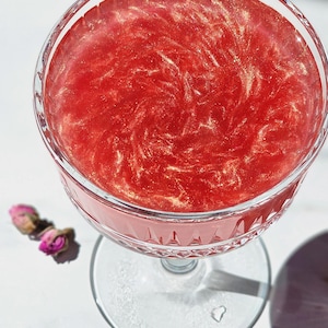 May include: A stemmed coupe glass containing a red cocktail with gold glitter swirls. The glass is on a white surface, with dried pink rose buds nearby. The drink's colour is a deep, rich red, creating an elegant aesthetic.