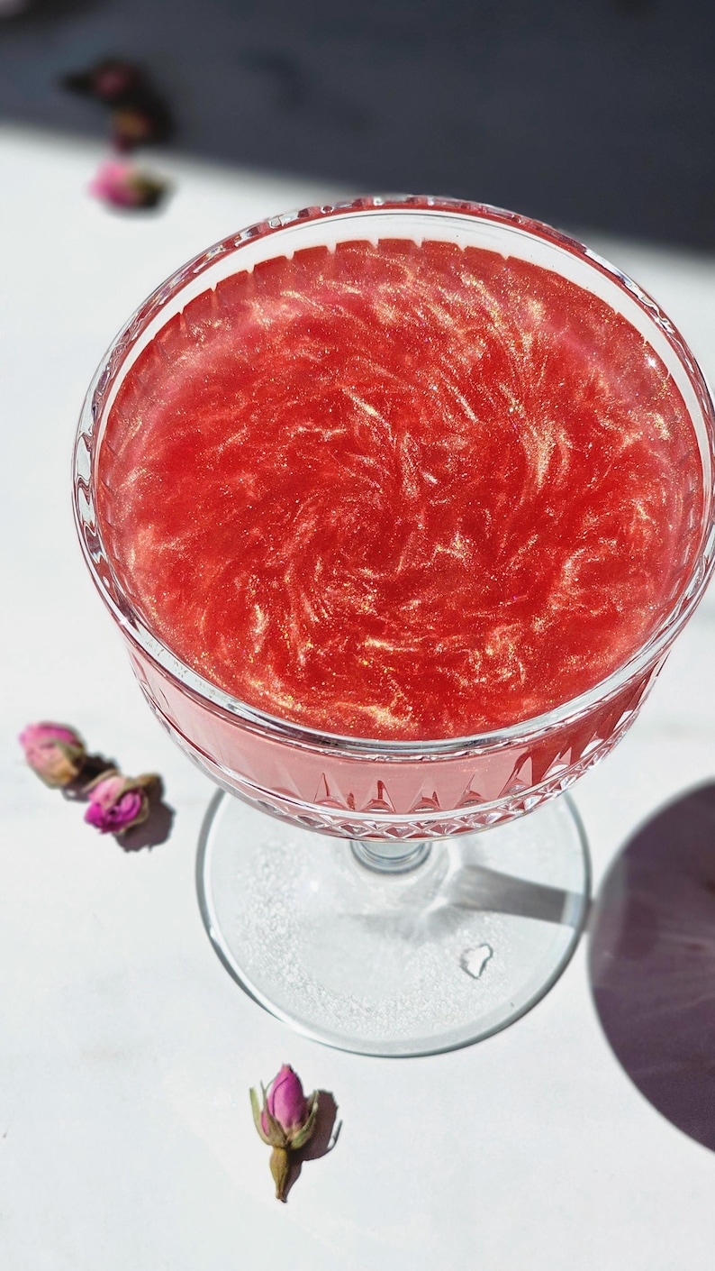 May include: A stemmed coupe glass containing a red cocktail with gold glitter swirls. The glass is on a white surface, with dried pink rose buds nearby. The drink's colour is a deep, rich red, creating an elegant aesthetic.