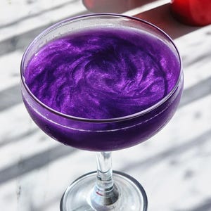 May include: A cocktail in a coupe glass, with a shimmering, swirling purple liquid. The drink reflects light, creating a glittery effect. The glass is on a marble surface, with red objects in the background.