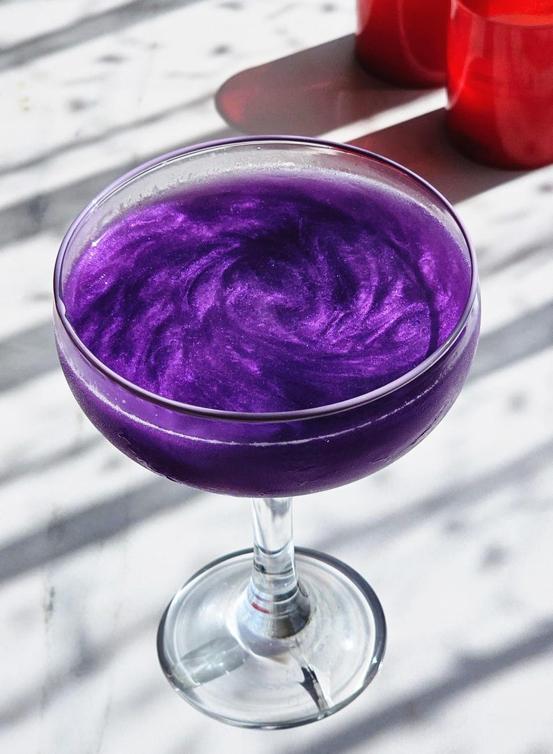 May include: A cocktail in a coupe glass, with a shimmering, swirling purple liquid. The drink reflects light, creating a glittery effect. The glass is on a marble surface, with red objects in the background.