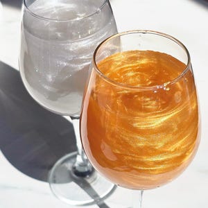 May include: Two wine glasses, one with a shimmering silver liquid and the other with a swirling gold liquid. The glasses are on a white surface, with shadows from sunlight. The glasses are made of glass.