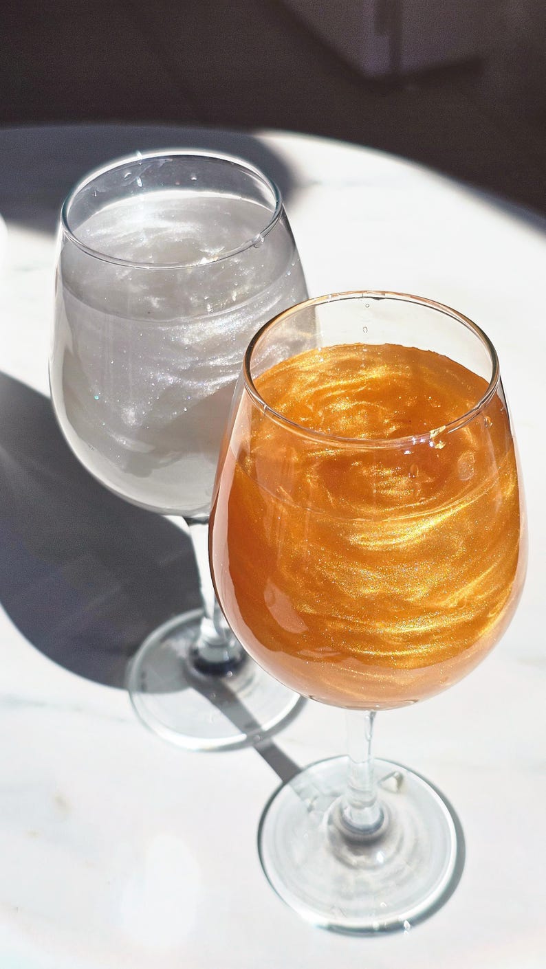 May include: Two wine glasses, one with a shimmering silver liquid and the other with a swirling gold liquid. The glasses are on a white surface, with shadows from sunlight. The glasses are made of glass.